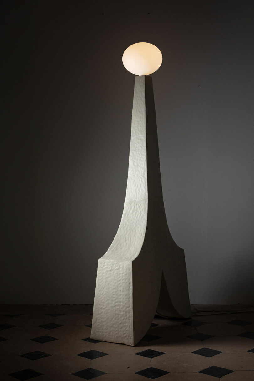 Tall sculptural lamp by Kelsie Rudolph features a textured, angular white base and a glowing spherical bulb, set against a plain, dimly lit background with a tiled floor.