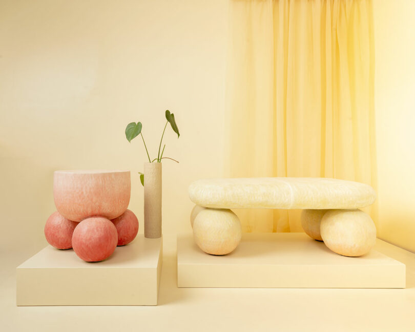 A round pink stool, a tall vase with two green leaves, and an oval yellow bench by Kelsie Rudolph sit on cream platforms against a pale yellow backdrop with sheer curtains.