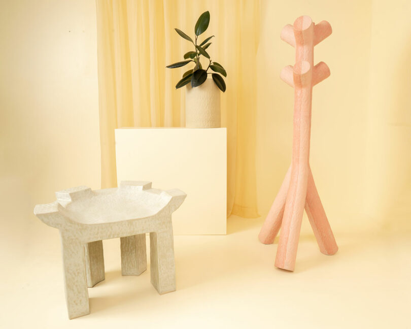 A textured white chair, a beige vase with a plant on a white block, and a pink sculptural coat rack by Kelsie Rudolph are arranged in a minimalist beige room.