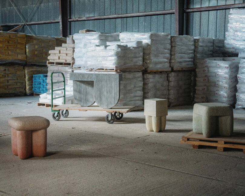 Industrial warehouse interior with stacked pallets of packaged goods and sculptural stools by Kelsie Rudolph arranged on the floor and a pallet.