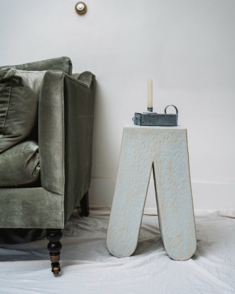 A green velvet sofa next to a white sculptural side table with a candle and a metal box on top, against a white wall with a plastic sheet on the floor
