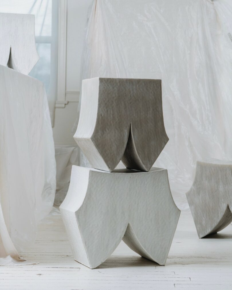 Two sculptural stools with angular, curved bases are stacked in a bright room with sheer drapes and white wooden floors