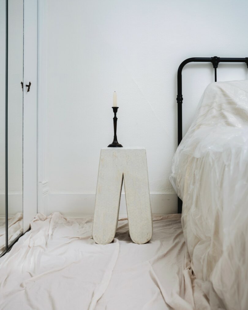 A minimalist bedroom with a black metal bed frame, a white bedside stool, a single candlestick with an unlit candle, and a draped white sheet on the floor