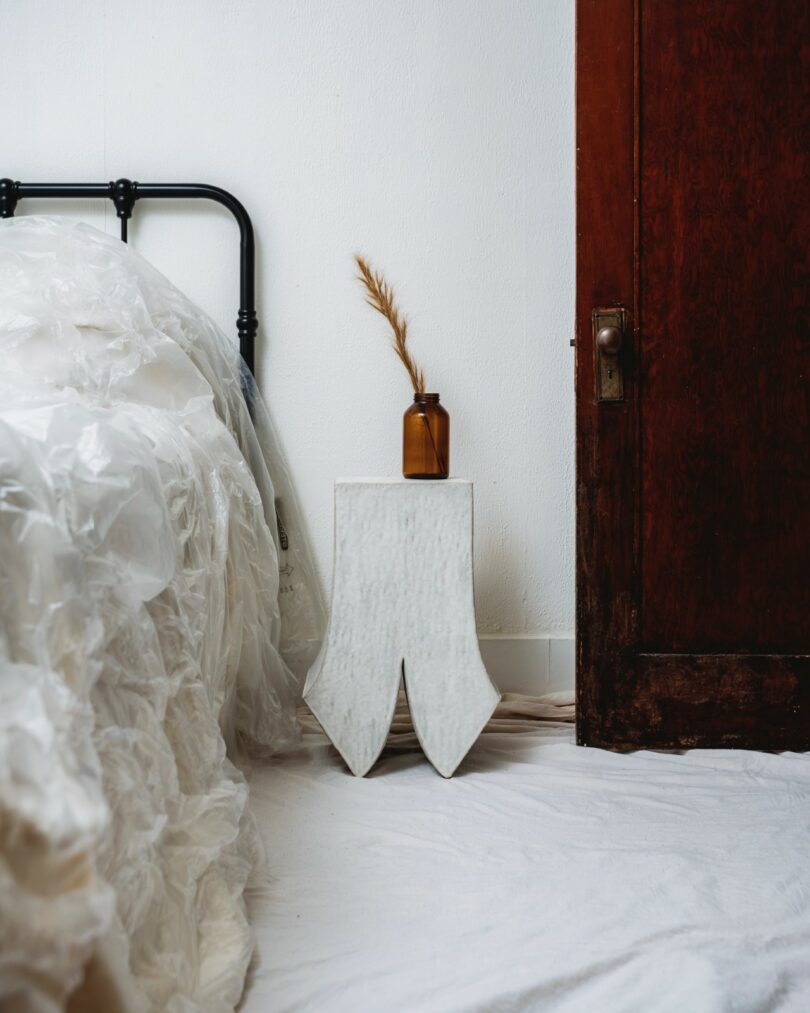 A brown glass bottle with dried grass sits on a white sculptural side table between a bed with a plastic cover and a dark wooden door