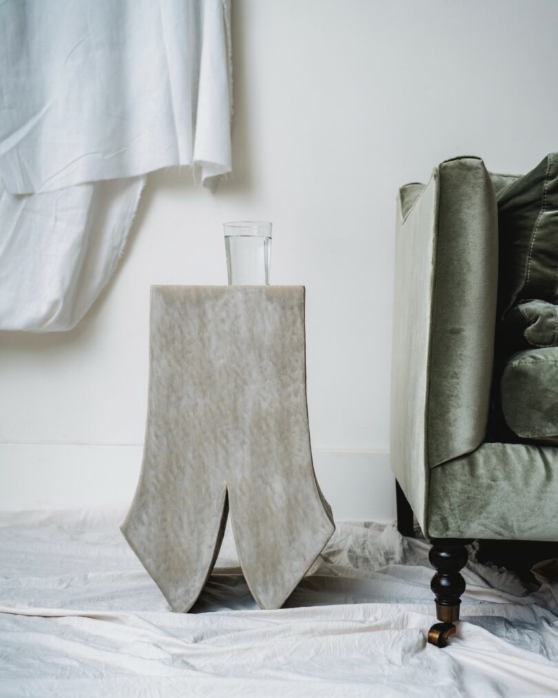 A glass of water sits on a beige, fabric-draped side table next to a green upholstered sofa, with a white sheet covering the floor and wall behind