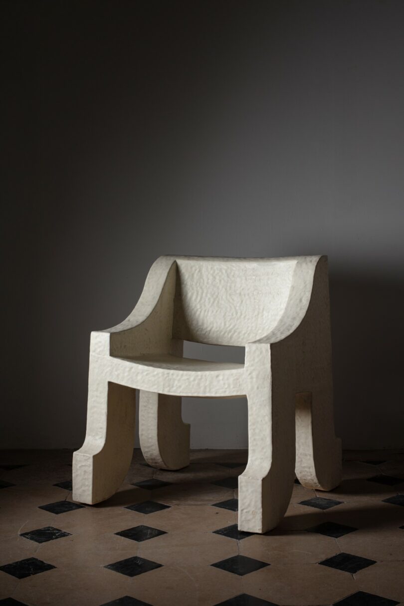 A sculptural, modern white chair with textured surface and curved backrest, set on a checkered tile floor against a plain gray wall