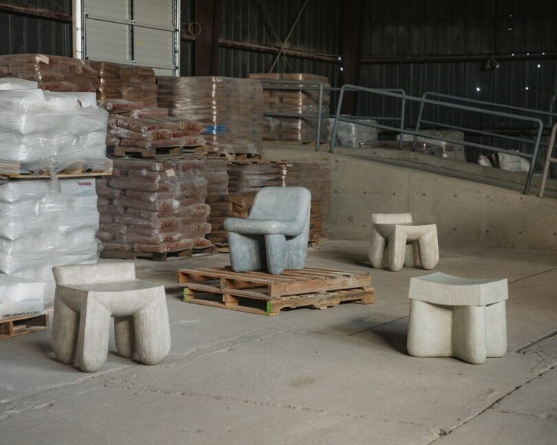 Four sculptural stone chairs are arranged on a concrete floor in a warehouse, surrounded by stacks of bagged goods and wooden pallets