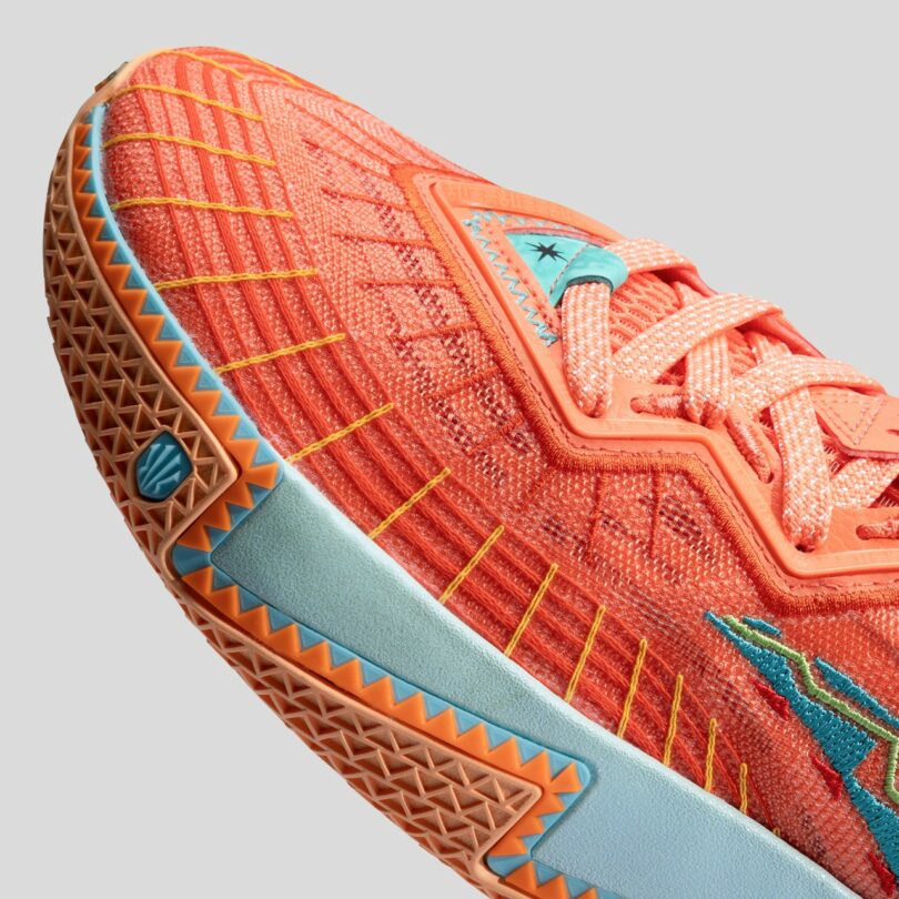 Close-up of an orange athletic shoe with blue and yellow accents, showing detailed stitching and textured sole against a plain gray background