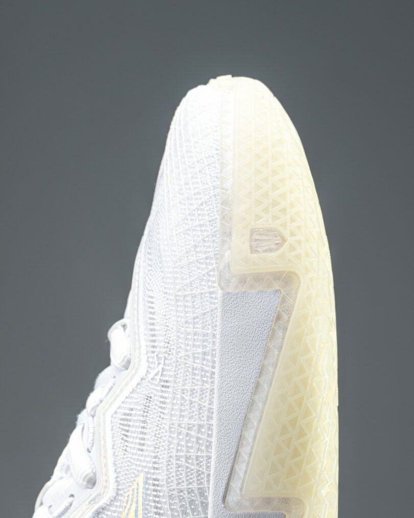 Close-up of a white athletic sneaker with textured and glossy detailing, showing the laces, tongue, and part of the upper design against a plain gray background.