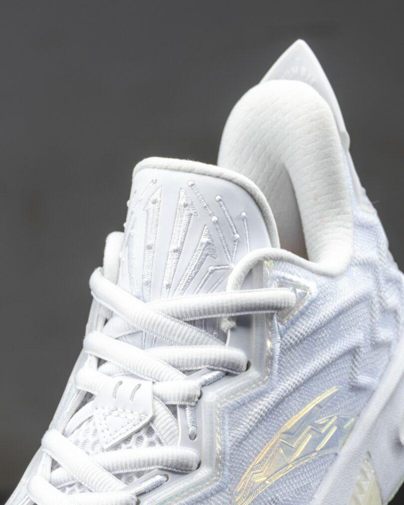 Close-up of a white athletic sneaker with textured and glossy detailing, showing the laces, tongue, and part of the upper design against a plain gray background