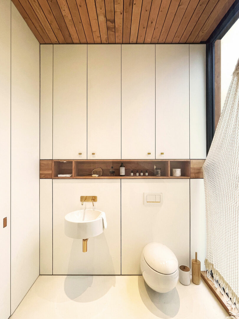 Modern minimalist bathroom with white cabinets, a round wall-mounted sink, an oval toilet, wooden accents, and a hammock chair partially visible on the right.