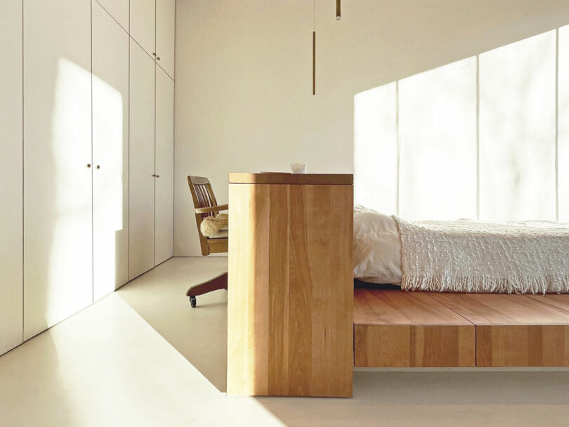 Minimalist bedroom with white walls, built-in closets, a wooden bed platform, a wooden chair, and sunlight streaming through large windows.