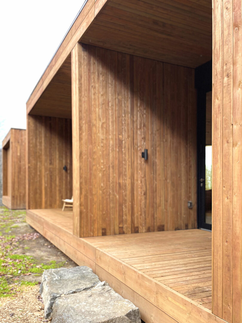 A modern building with vertical wooden siding and a covered porch, featuring a single chair and large stones beside the wooden deck.