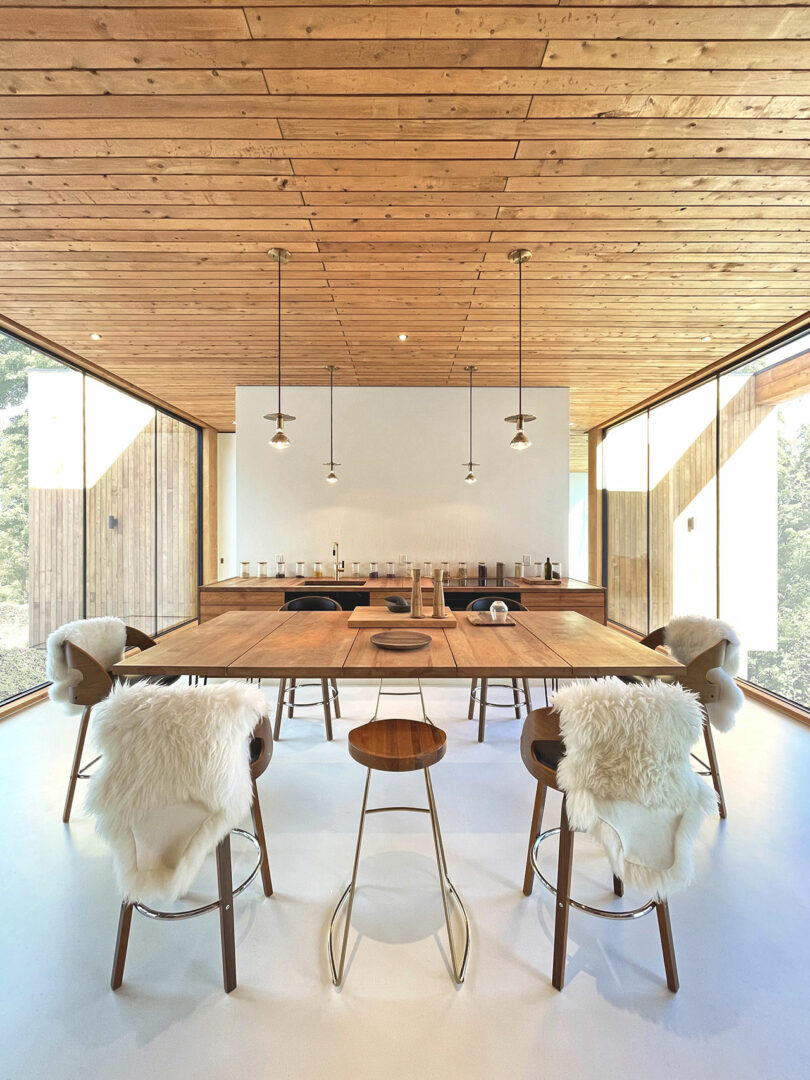 A modern dining room with a large wooden table, minimalist decor, pendant lights, and floor-to-ceiling windows; chairs have white fur throws draped over them.