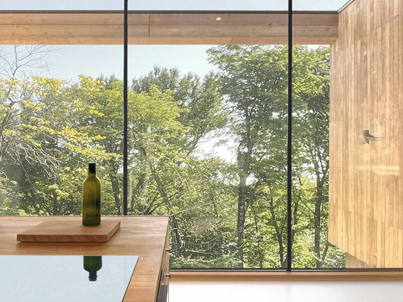 A green glass bottle on a wooden cutting board sits on a counter in front of large floor-to-ceiling windows overlooking leafy trees.