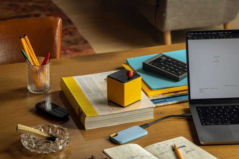 A desk with a laptop, notebooks, a glass of pencils, an external drive, a remote control, books, an ashtray, and a small yellow device with a red button.