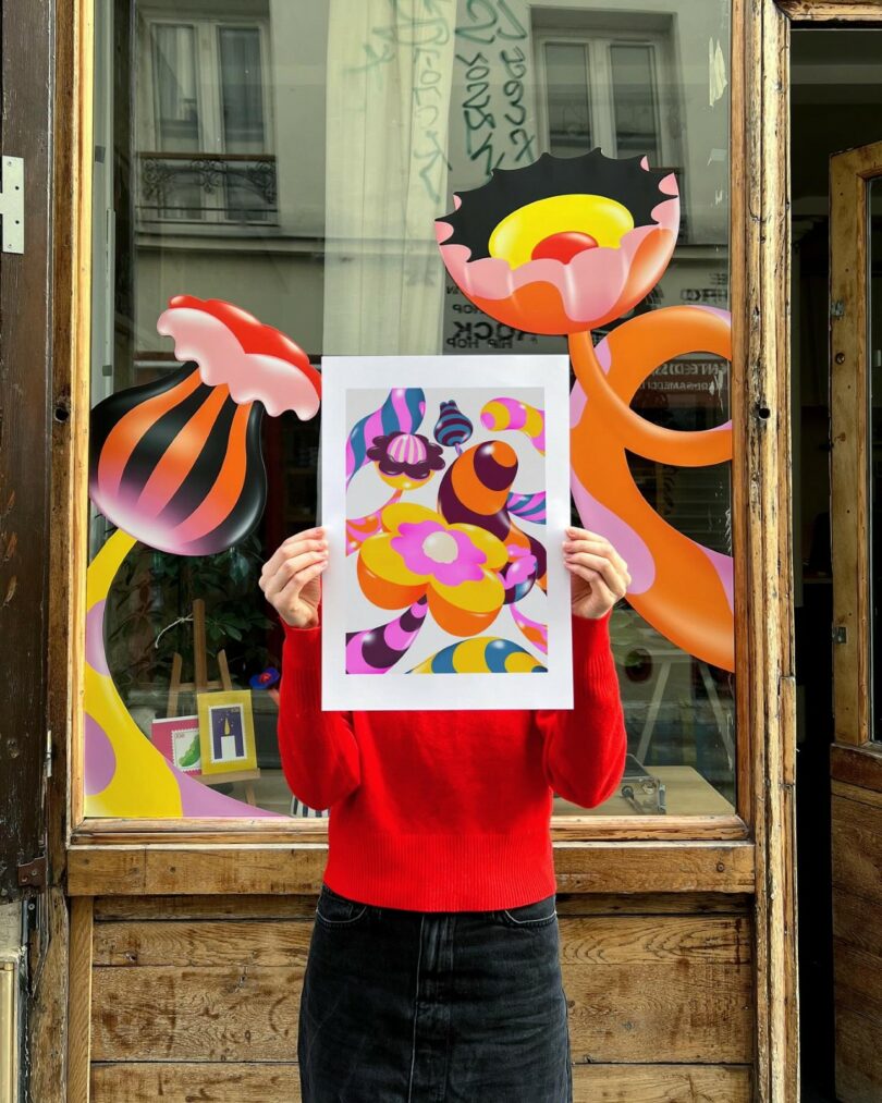 A person in a red sweater holds up a colorful abstract art print, aligning it with similar art painted on a storefront window behind them