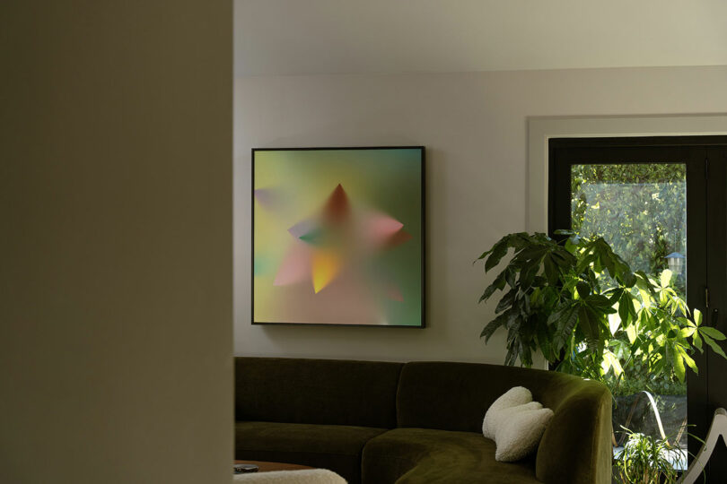 A minimalist living room with a curved green sofa, a large leafy plant, and a wall-mounted abstract artwork featuring soft, colorful shapes.