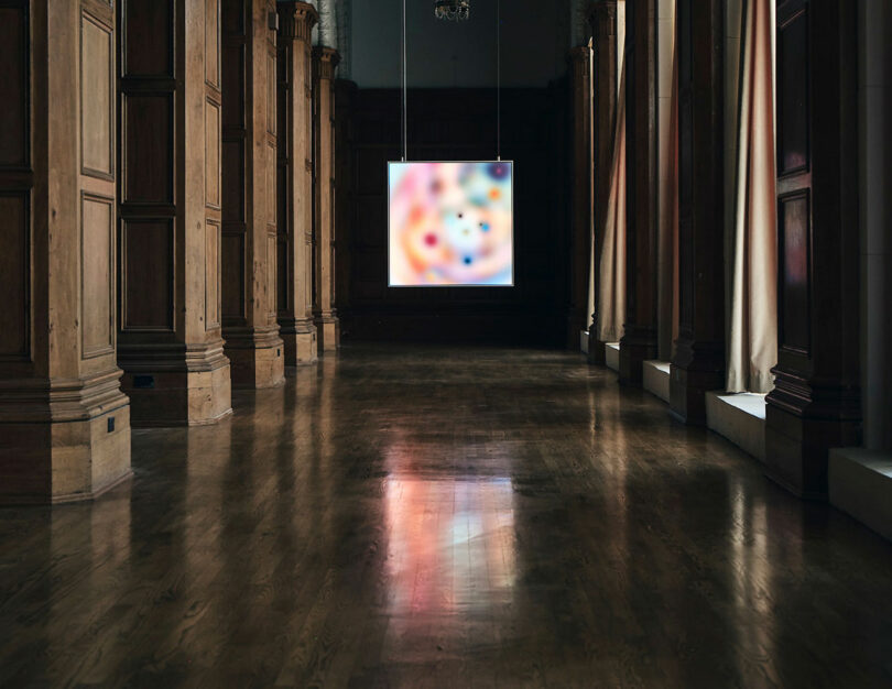 A large, blurred, colorful artwork is suspended in the center of an ornate, wooden-paneled room with tall columns and polished floors.