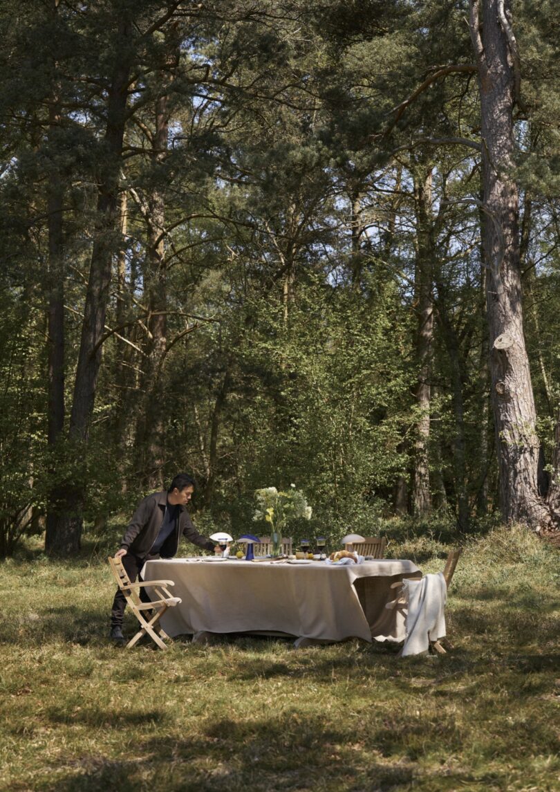 A person stands beside a table set for a meal with chairs, dishes, and flowers, situated in a forest clearing surrounded by tall trees