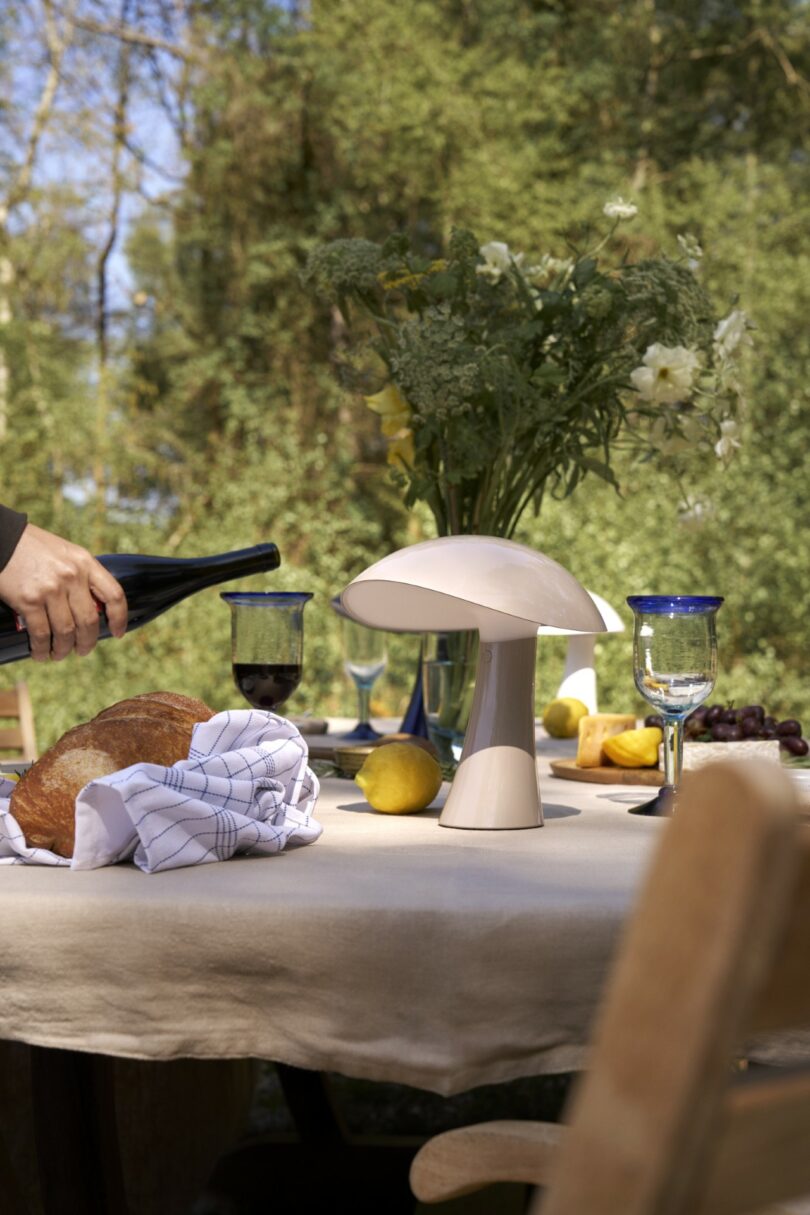 A table set outdoors with bread, wine, fruit, glasses, a vase of flowers, and a mushroom-shaped lamp, with a person pouring wine