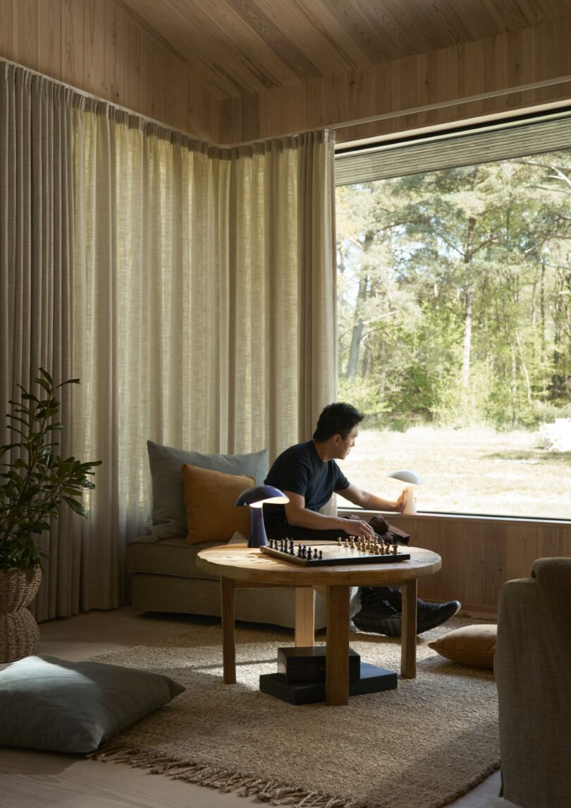 A person sits in a cozy living room by a large window, looking outside. A round wooden table with a chessboard and lamp is in the foreground