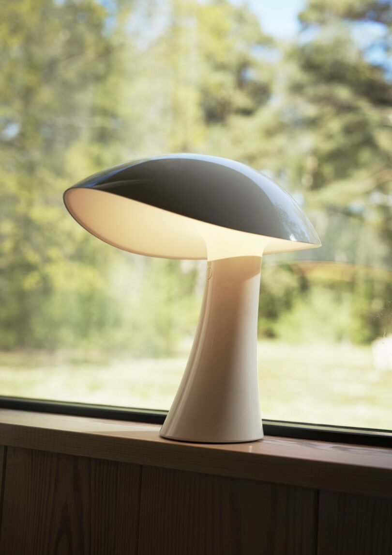 Modern white mushroom-shaped table lamp on a wooden windowsill, illuminated, with a blurred view of trees and greenery outside