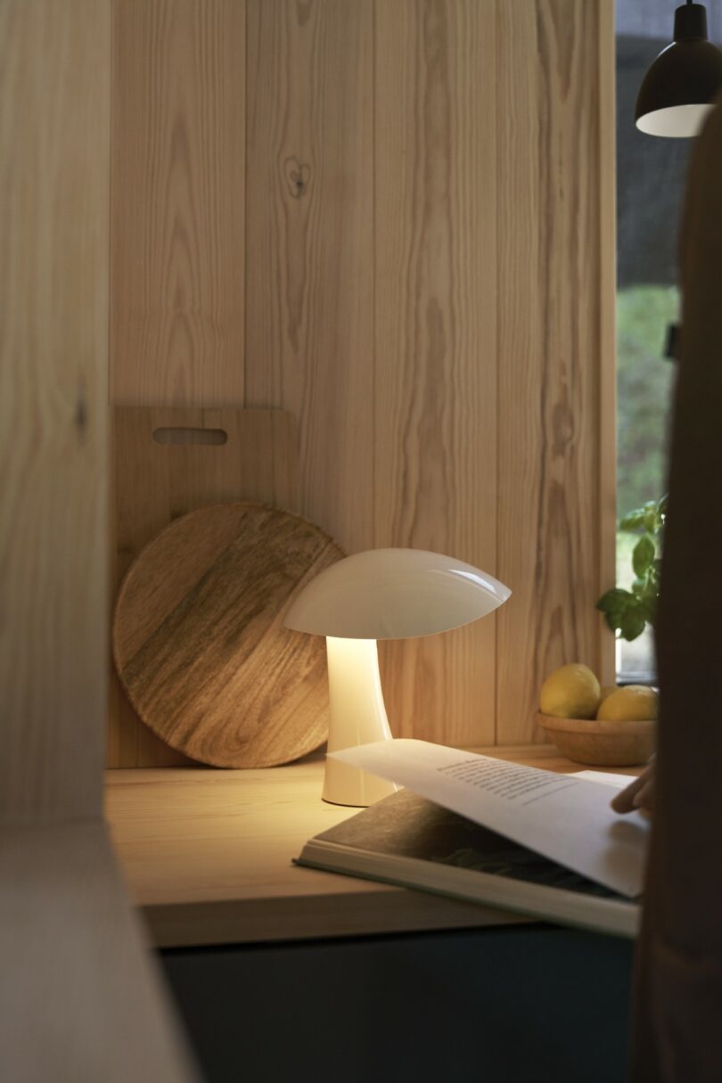 A small white mushroom-shaped lamp on a wooden desk, with a round cutting board, lemons, and a hand flipping through a book nearby
