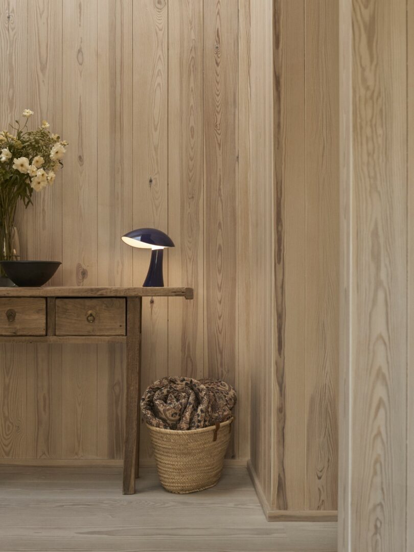 A wooden console table with drawers sits against light wood-paneled walls, holding a black bowl, a vase of flowers, and a blue table lamp. A woven basket with blankets is below