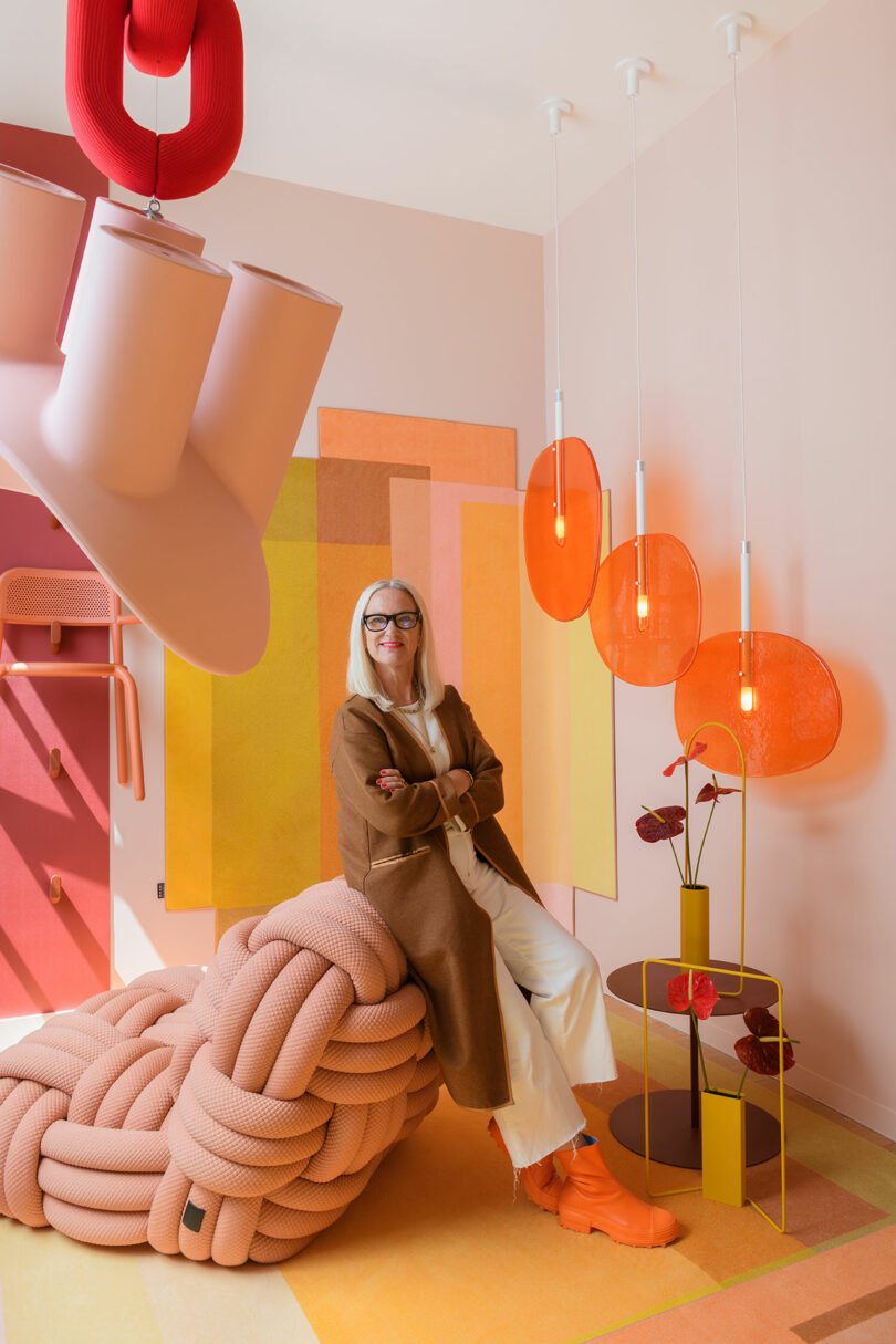 A woman in a brown coat and orange shoes sits on a large pink knotted seat in a colorful room with orange lamps, geometric wall art, and modern decor.