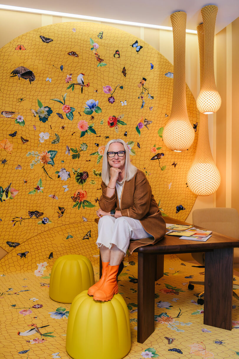 A woman with long white hair and glasses sits on a yellow stool in a colorful room with floral and butterfly designs on the walls and floor, next to a wooden table and hanging lamps.