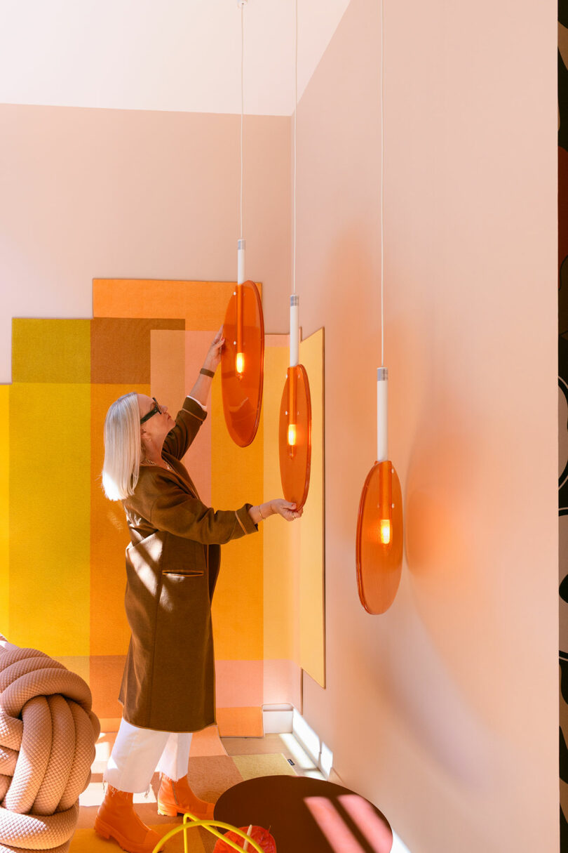 A woman with gray hair adjusts one of three orange pendant lights in a modern, brightly colored room with geometric wall panels and a large woven rope decor.
