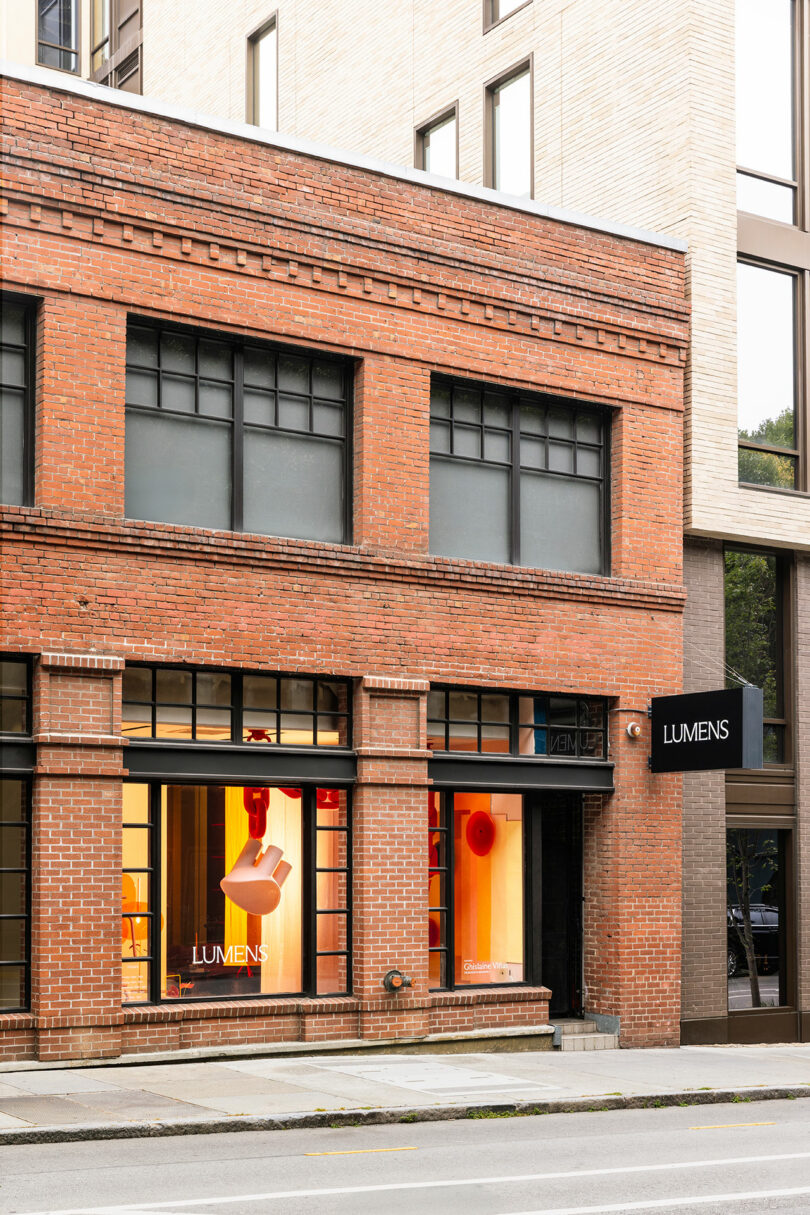 A brick building with large windows displays modern lighting fixtures. A black sign and window read "LUMENS." The street and part of another building are visible.
