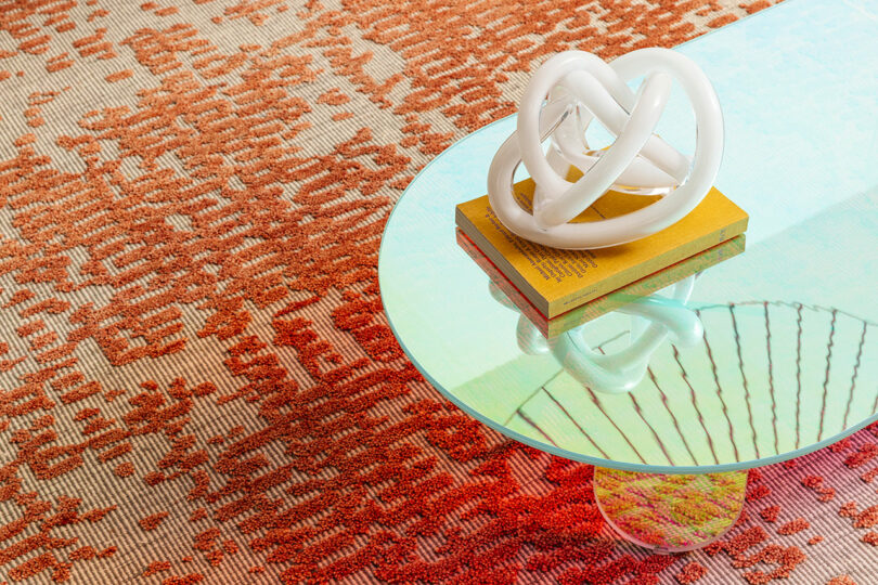 A glass coffee table on a red patterned rug, with a yellow book and a white knotted sculpture on top. The table surface reflects the sculpture and rug.