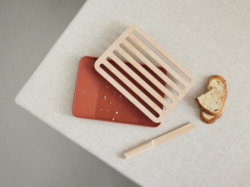 A wooden slotted breadboard on a red tray sits on a beige tablecloth, with crumbs, a bread knife, and two slices of bread nearby