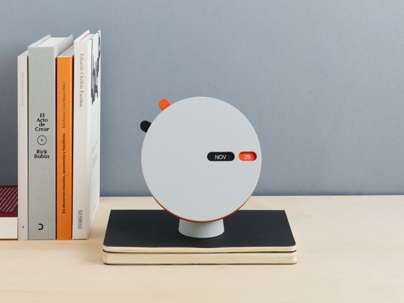 A round, minimalist desk calendar displaying "Nov 29" sits on a closed notebook beside upright books on a light wood desk against a light gray wall