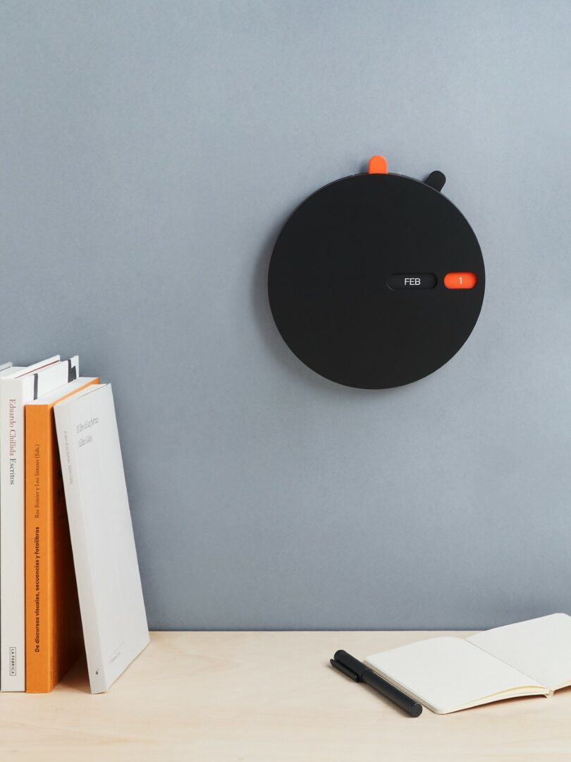 A round, black wall calendar with sliding indicators displays "FEB" and "1." It is mounted above a desk with books, an open notebook, and a pen