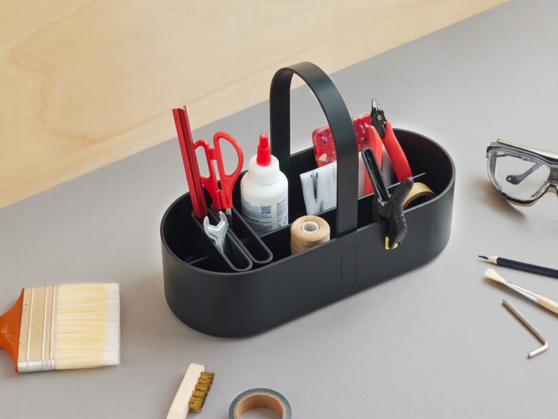 A black caddy holding scissors, pens, glue, tape, wire cutters, a clamp, and other tools sits on a table with additional craft supplies scattered nearby