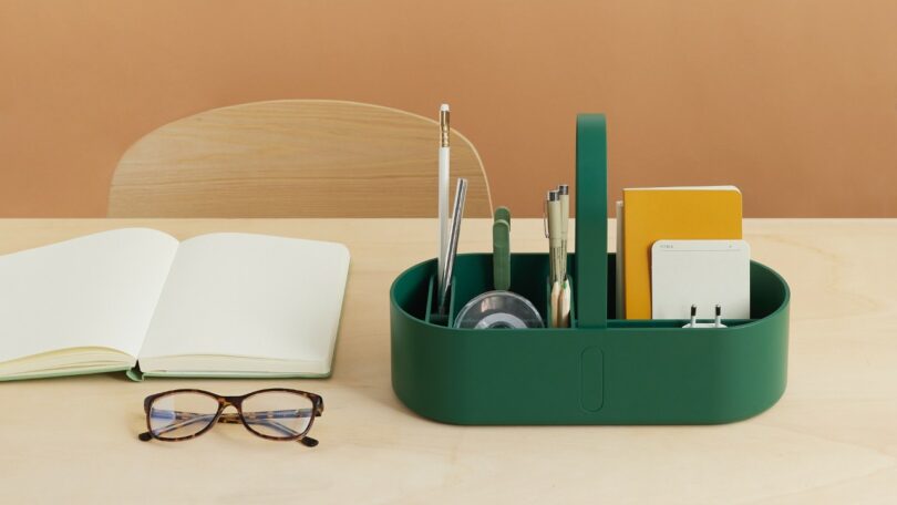 A green desk organizer holds pens, tape, scissors, and notebooks on a light wood table next to an open notebook and a pair of eyeglasses