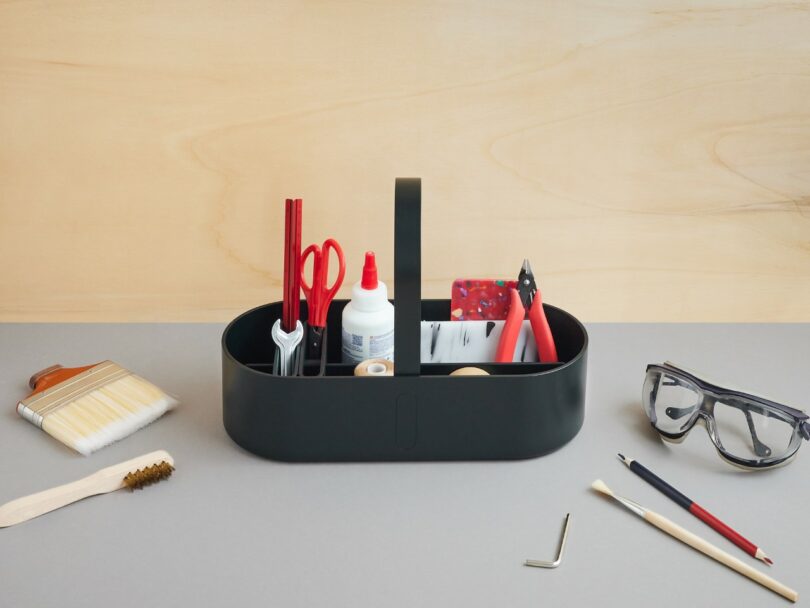 A black organizer tray holds art and craft supplies, including glue, scissors, pliers, pencils, and brushes; goggles and tools are placed on the table nearby