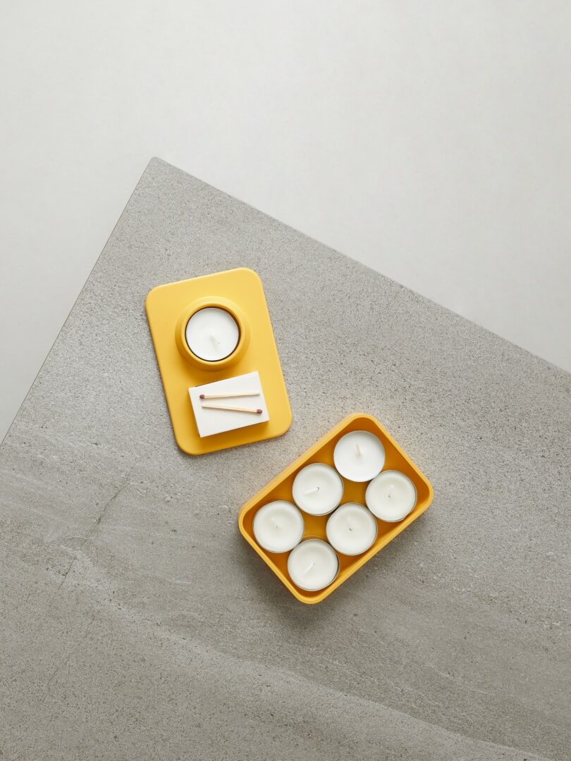 A yellow tray with a candle and two matches, and a yellow container holding six tealight candles, placed on a gray textured surface