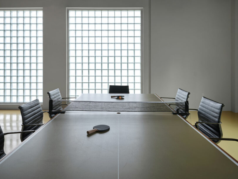 A conference table with a ping pong net, four office chairs, two paddles, and a ball, set in a modern room with large glass block windows.