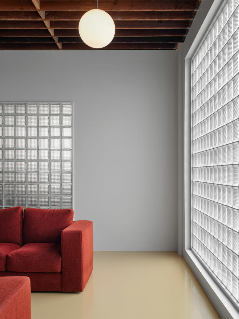 A modern room with a red sofa, large glass block windows, a pale floor, a white wall, and an exposed wooden beam ceiling with a round ceiling light.