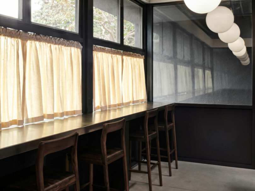Long wooden counter with several wooden chairs lined up by large windows with beige curtains; round ceiling lights hang above.
