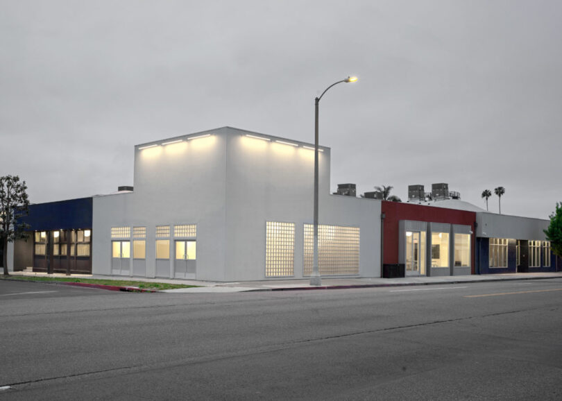 A modern, single-story commercial building with white, blue, and red exterior walls, lit windows, and a streetlamp on a quiet, empty street at dusk.