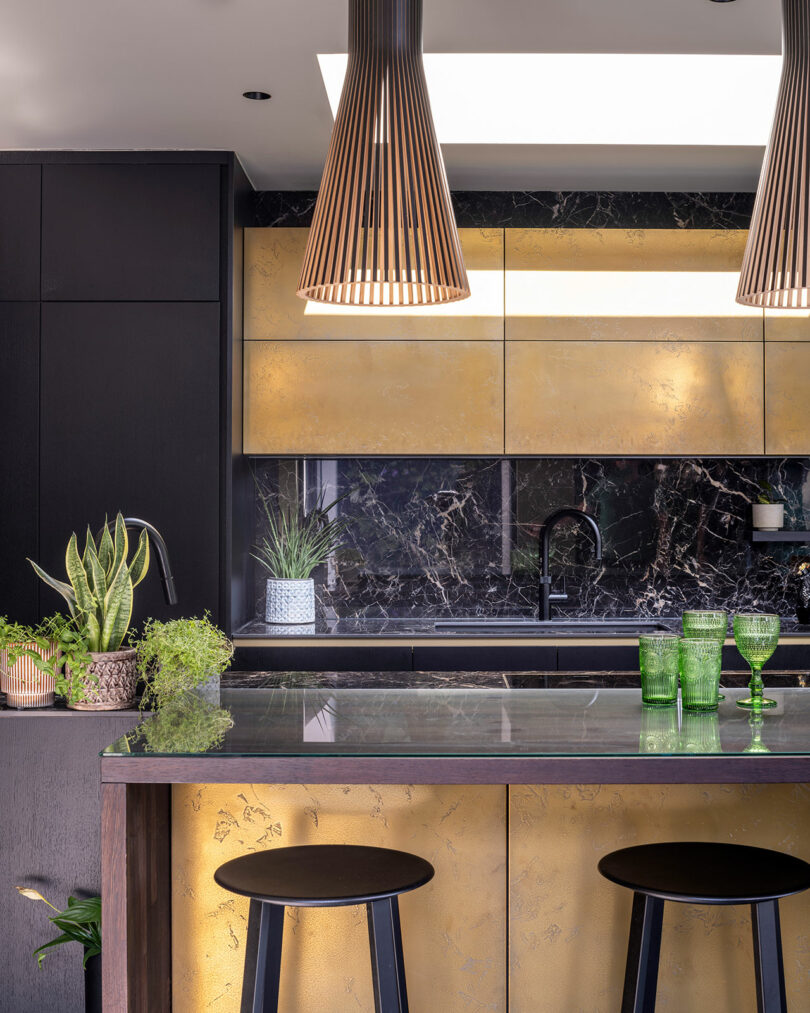 Modern kitchen with gold cabinets, black marble backsplash, green plants, two black stools, and green glassware on the counter under pendant lights.