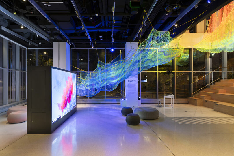 A modern gallery space with a large digital screen, round seating, and a colorful suspended net-like installation under bright lighting.