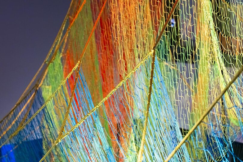 Colorful, translucent nets with wavy patterns are suspended diagonally, overlapping each other in shades of orange, yellow, green, and blue against a neutral background.