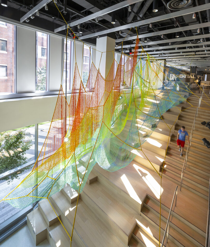 A large, colorful net installation is suspended above wooden steps inside a modern building with large windows and exposed ceiling beams.