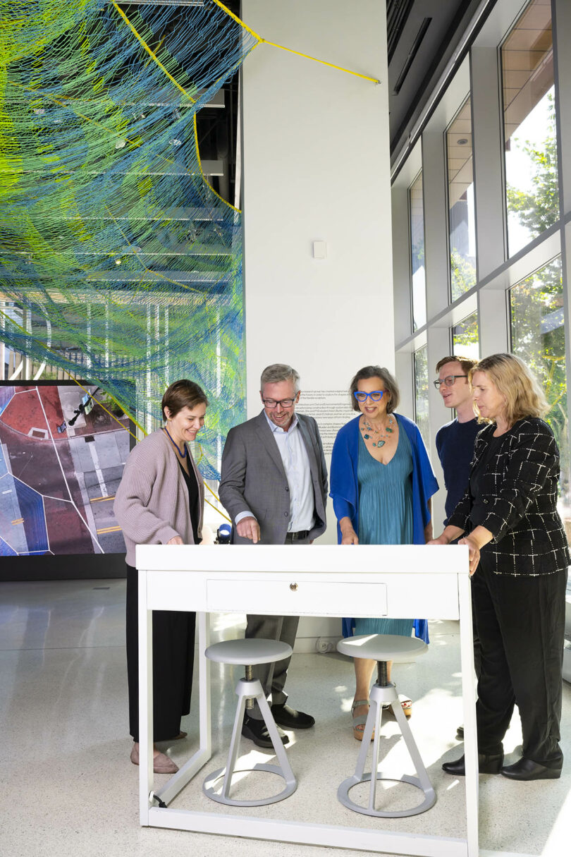 Five adults stand around a white table with two stools in a modern, well-lit room featuring colorful suspended artwork and large windows.
